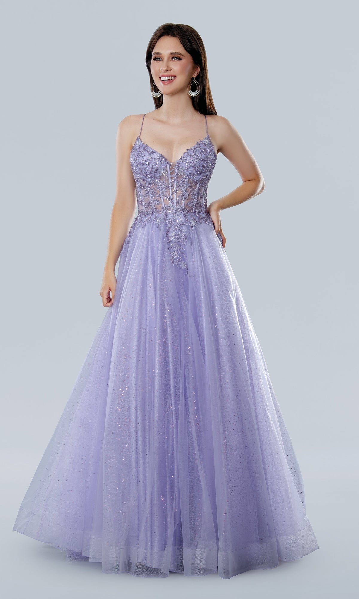 A woman smiles against a plain background in the Stella Couture 25140 Glitter Prom Ball Gown—a floor-length, sleeveless lavender dress with a fitted V-neck corset bodice featuring sequin lace embroidery and a full tulle skirt.